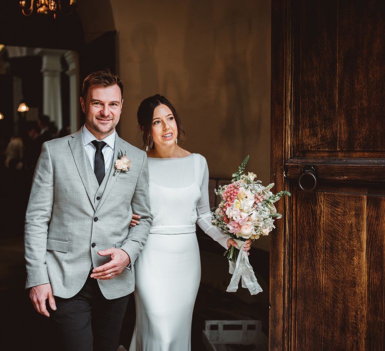 Groom in in black suit trousers, grey blazer and light pink rose boutonniere linking arms with bride in long satin wedding dress with bridal cover up semi sheer crop top holding tropical wedding bouquet with pink garden roses, foliage and baby's-breath 