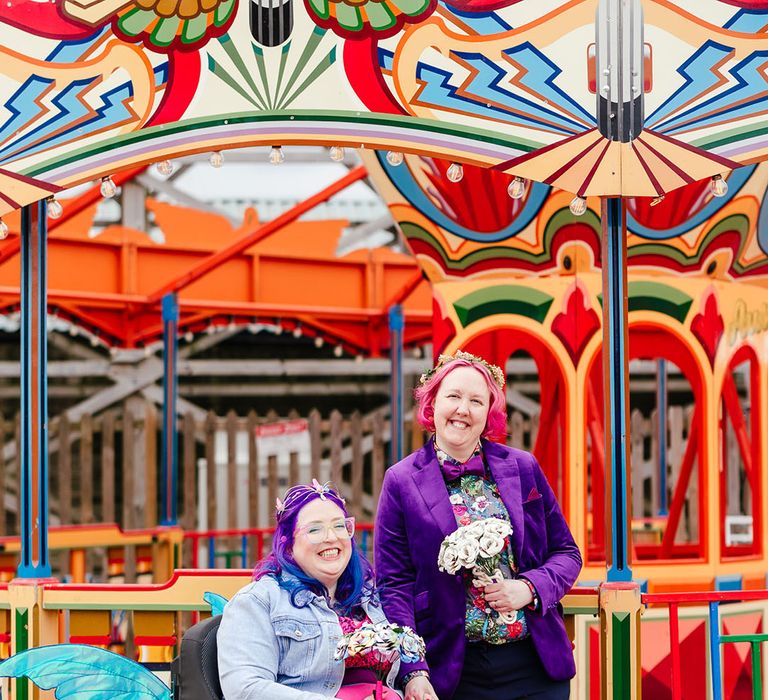 Bride in wheelchair with sparkly blue fairy wings hung on wearing personalised denim jacket, pink wedding dress and blue fishnet tights with bride in purple velvet blazer and black suit trousers with purple bowtie, pink patterned pocket square and colourful patterned shirt