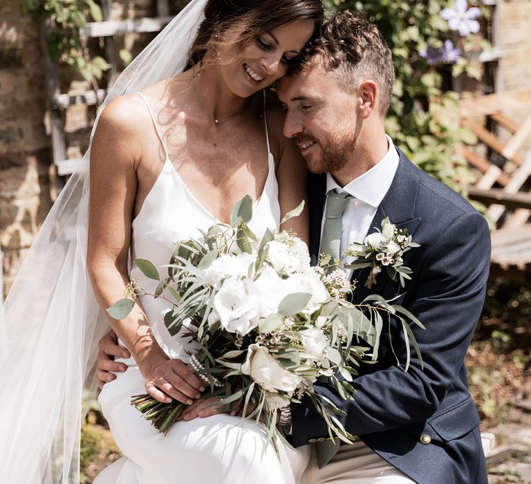 Bride holds white floral bouquet and Grace Loves Lace wedding dress whilst sat beside her groom in double breasted blazer 