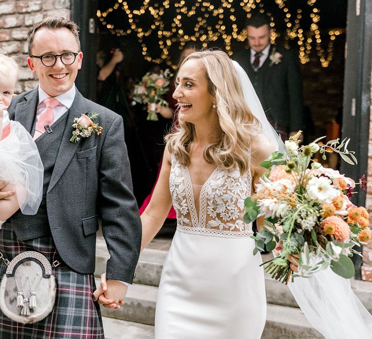 Bride in Mikaella Bridal wedding dress with appliqué bodice walks alongside her groom in kilt 