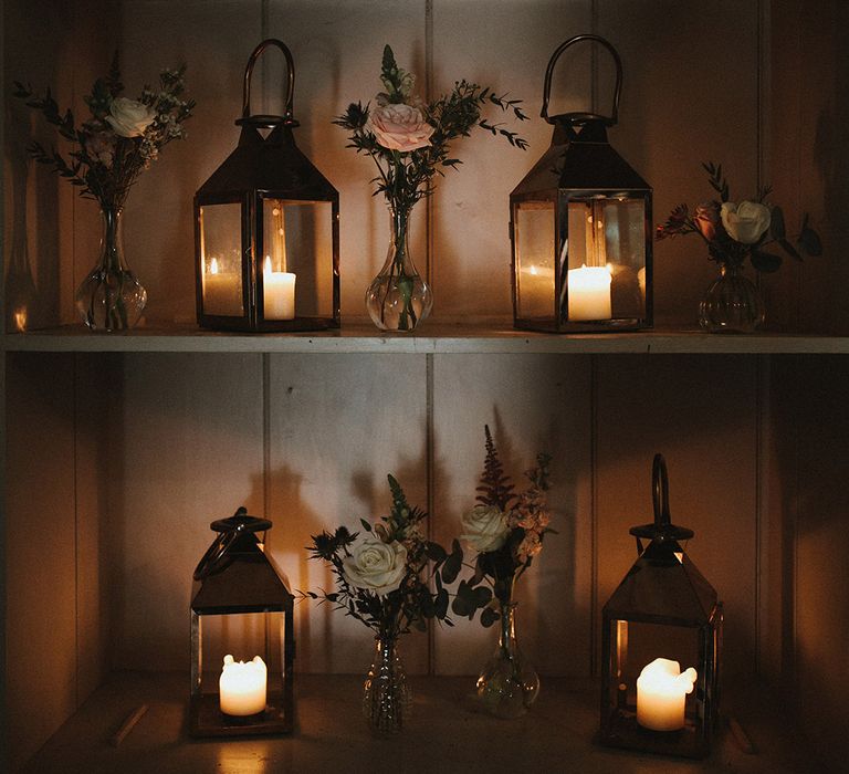 White pillar candles in glass lanterns on a shelf with bud vases full of pink and white roses 