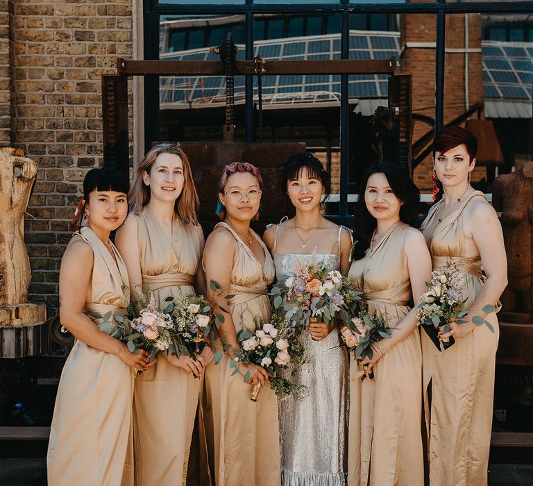 Bride stands with her bridesmaids in gold bridesmaid dresses with halter necks and homemade bouquets 