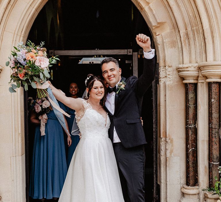 Bride in flower patterned wedding dress puts her arm in the air with the groom to celebrate their wedding with groom in black tie 