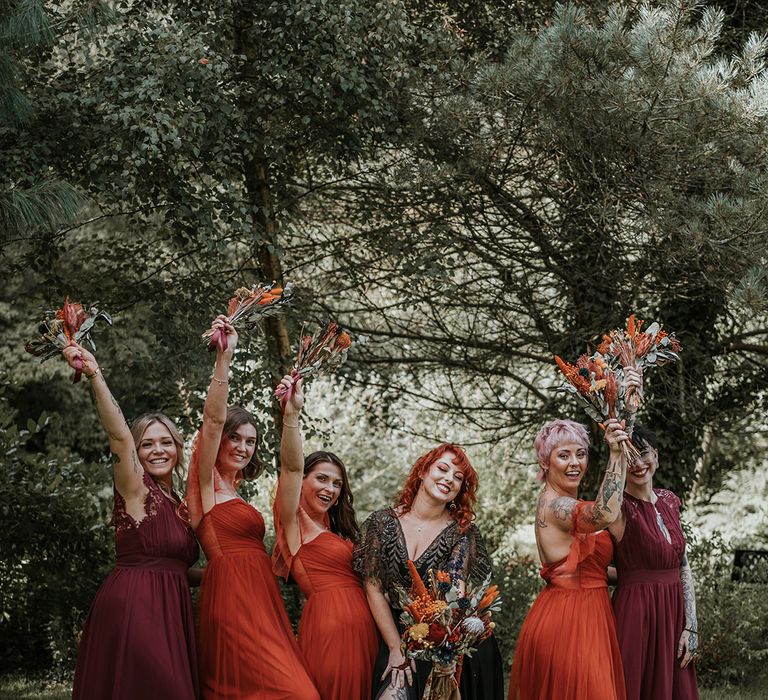 Bride in black dress for wedding in Devon standing with bridesmaids in mismatched burgundy and burnt orange bridesmaid dresses holding burnt orange dried flower bouquets 