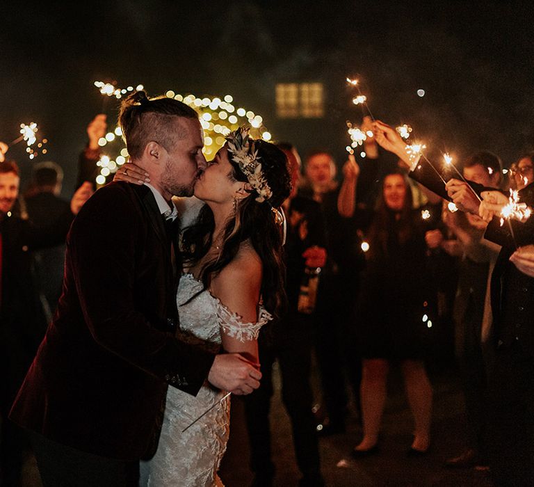 The bride and groom share a kiss at their boho autumnal wedding with the wedding guests waving sparklers for the sparkler send off 