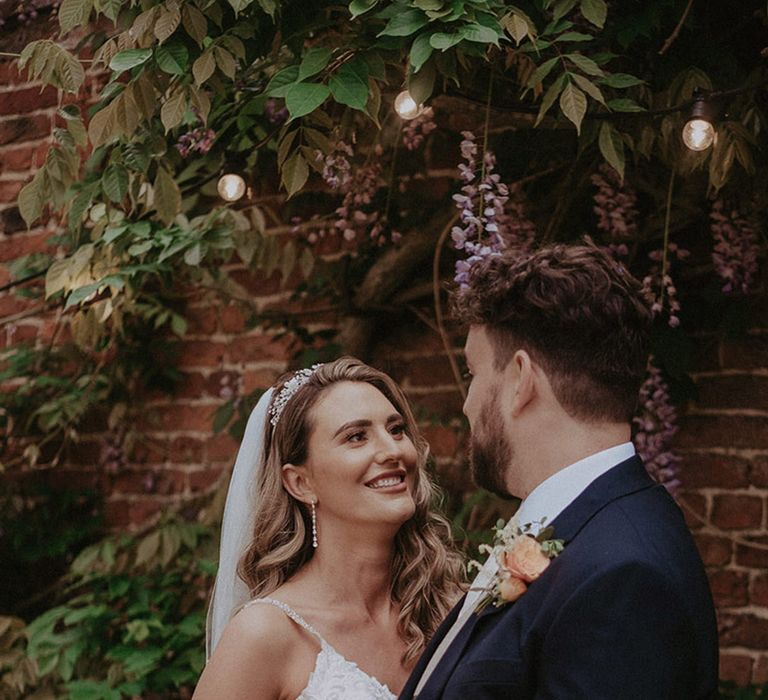 The bride smiles and gazes at the groom wearing a lace sparkly wedding dress with a peach and white rose bouquet with the groom in a navy suit jacket