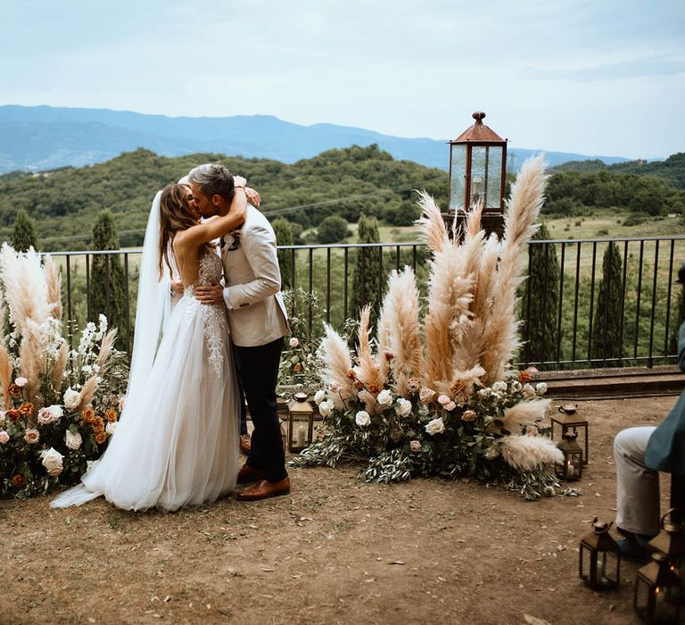 Bride and groom kiss at altar surrounded by pampas grass and rose decor in the Tuscan hills at destination wedding ceremony