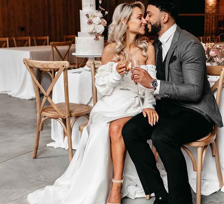 Groom in grey blazer, black waistcoat and navy polkadot tie having champagne with bride in off the shoulder long sleeve wedding dress with slight cowl neck and front leg slit 