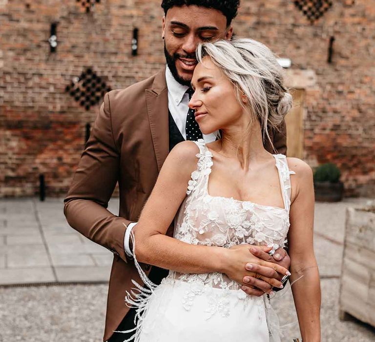 Bride in Nori Ivy corset top sheer floral 3d appliqué wedding dress with fringe overlay with groom in mocha brown blazer, black waistcoat, polka dot tie and pocket square and dried blush boutonniere on the grounds of Lodge Farm wedding venue 