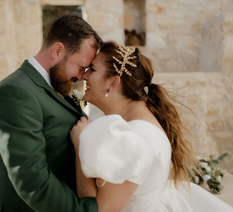 Bride in white satin dress and gold leaf bridal crown touches noses with groom in dark green groom suit at private villa wedding in Greece