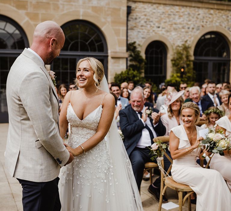 The bride wears a detailed applique wedding dress holding hands with the groom in a light grey suit jacket 