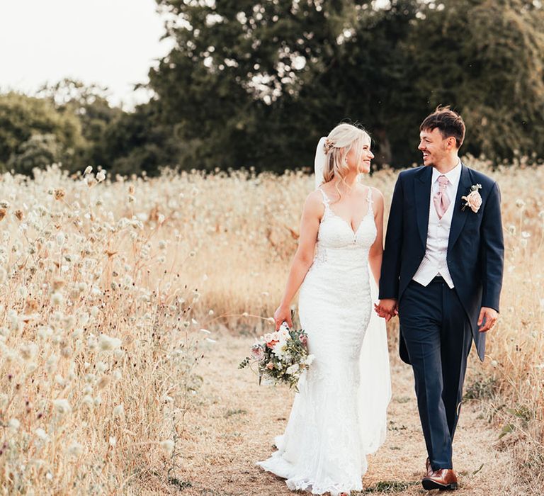 Bride walking along the grounds at Rackley Chiltern Hills barn wedding venue hand in hand with the groom in a navy suit 