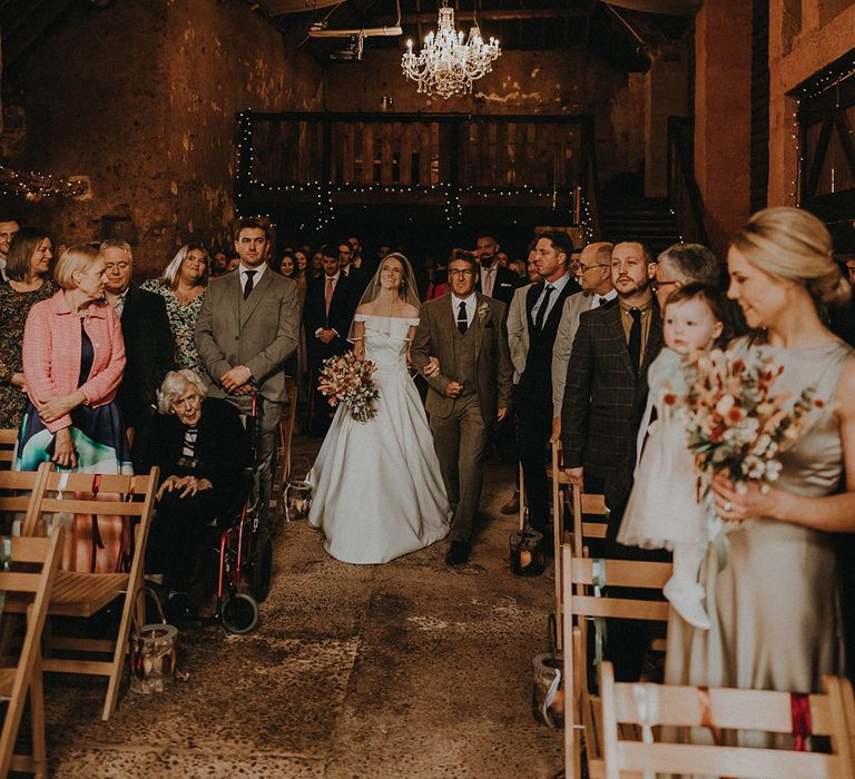 Bride in off the shoulder wedding dress walking down the aisle with the father of the bride at rustic romance theme barn wedding 