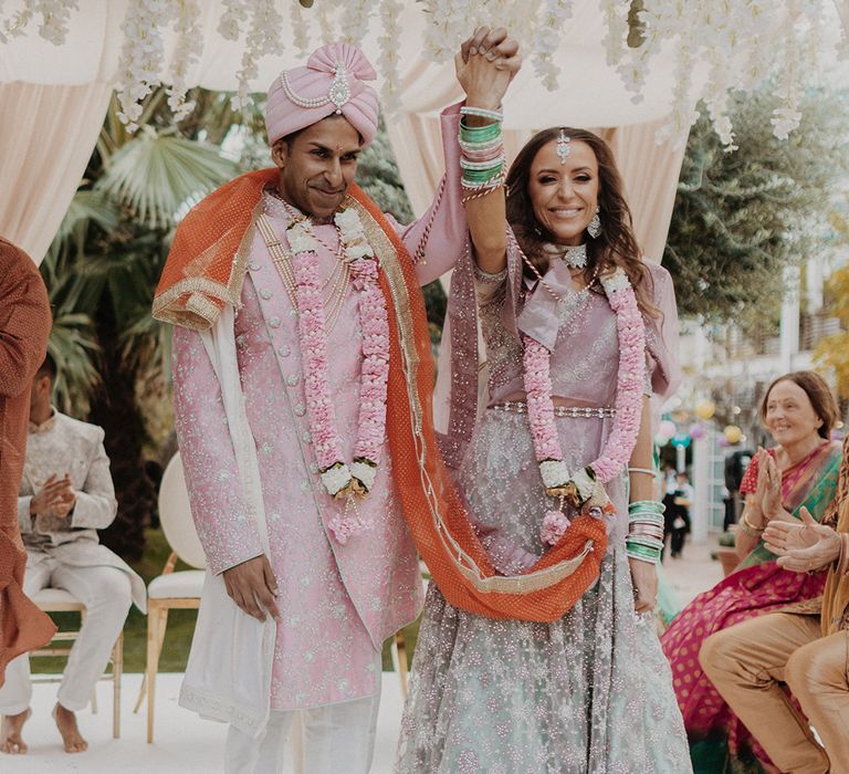 Groom in light pink sherwani, light pink turban and cream trousers raising hand with bride in mint green and dusky pink ombre embellished lehenga 