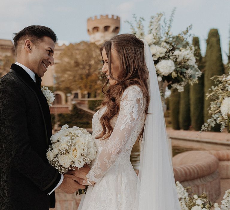 Bride in lace long sleeve Pronovias wedding dress and church-length veil holding white rose and dried flower bridal bouquet posing with groom in black tuxedo with white flower boutonniere 