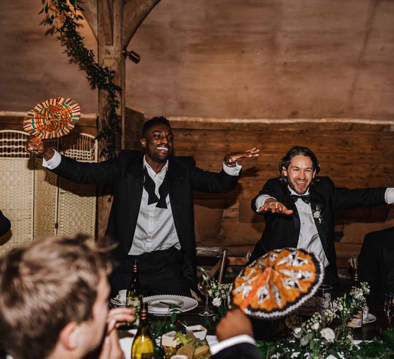 Wedding guests in classic black suits dancing and laughing at Lains Barn holding Ghanaian printed hand fan favours during wedding breakfast 