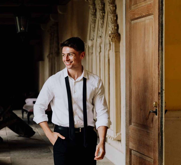 Groom walking through Euridge Manor wearing crisp white shirt, black suit trousers and black tie around his neck 