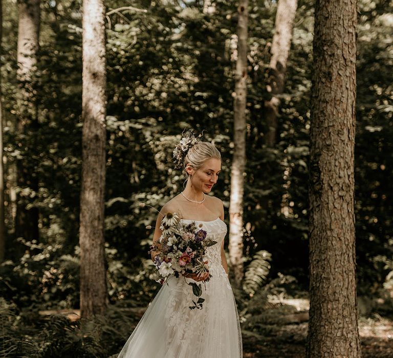 Bride in floral strapless wedding dress for woodland wedding with her hair in a chic updo decorated with purple and white wedding flowers 