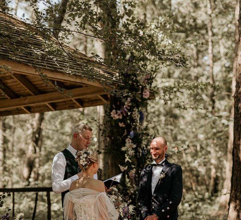 Bride in strapless wedding dress with tulle detachable sleeve standing facing the groom for outdoor woodland wedding ceremony 