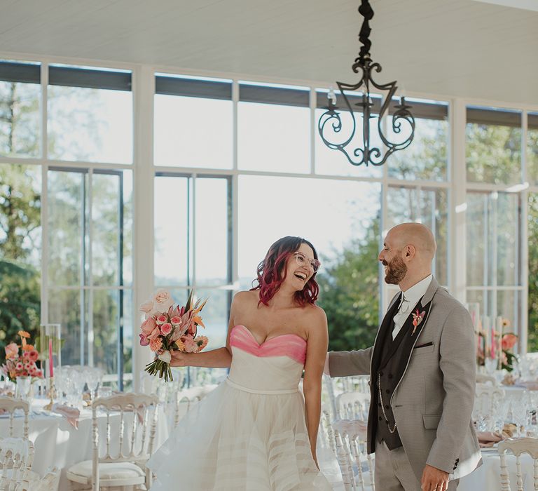 bride with glasses in a strapless wedding dress with layered skirt smiling at her groom in a grey wedding suit at their italian villa wedding reception