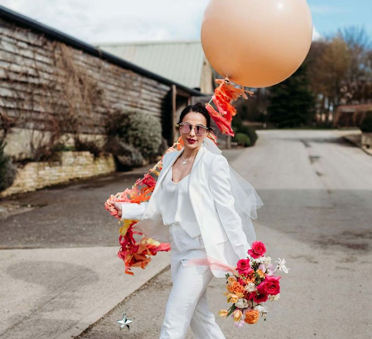 Bride in white bridal suit with closed toe bright green wedding heels holding mixed seasonal floral bouquet and large peach balloon with wedding streamers attached outside of Merriscourt wedding venue