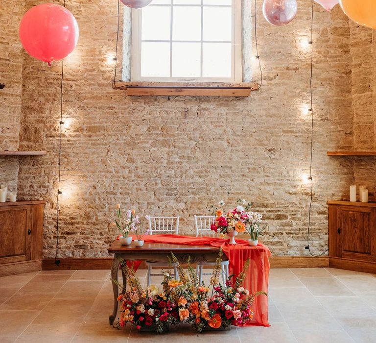 Merriscourt wedding venue reception room with metallic pink, white, orange and yellow balloons, wedding fairy lights decorations and wooden table with orange table runner and mixed seasonal flower arrangements 