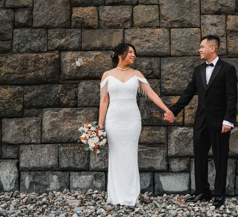 Bride in white beaded and sequined slip wedding dress holding hands with groom in classic black tux at Burmese wedding