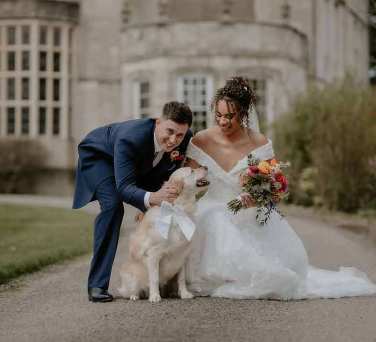 Bride in Suzanne Neville off shoulder 3D floral applique wedding dress posing with groom in navy groom morning suit with grey ties and blush rose boutonniere and golden retriever with white ribbon around his neck