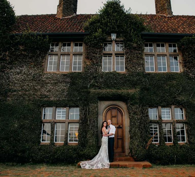 Bride in lace sleeveless wedding dress with puddle train embracing groom in white shirt, black tie and black suit trousers outside of Giraffe Manor wedding venue Kenya