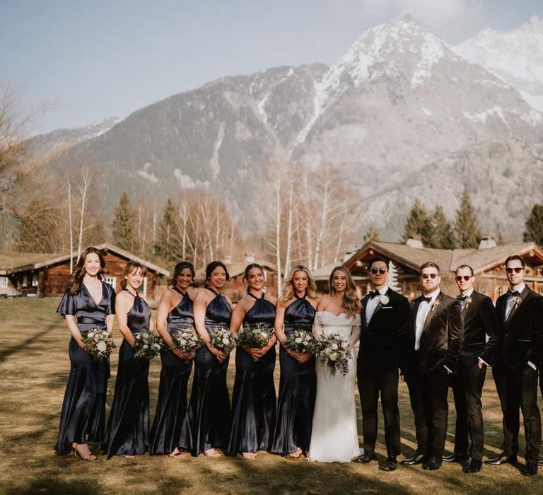 Groom and groomsmen in classic black tuxedos, black bowties and white garden rose boutonnieres standing with bride in off shoulder sparkly mermaid wedding dress and bridesmaids in satin halterneck navy bridesmaid dresses  