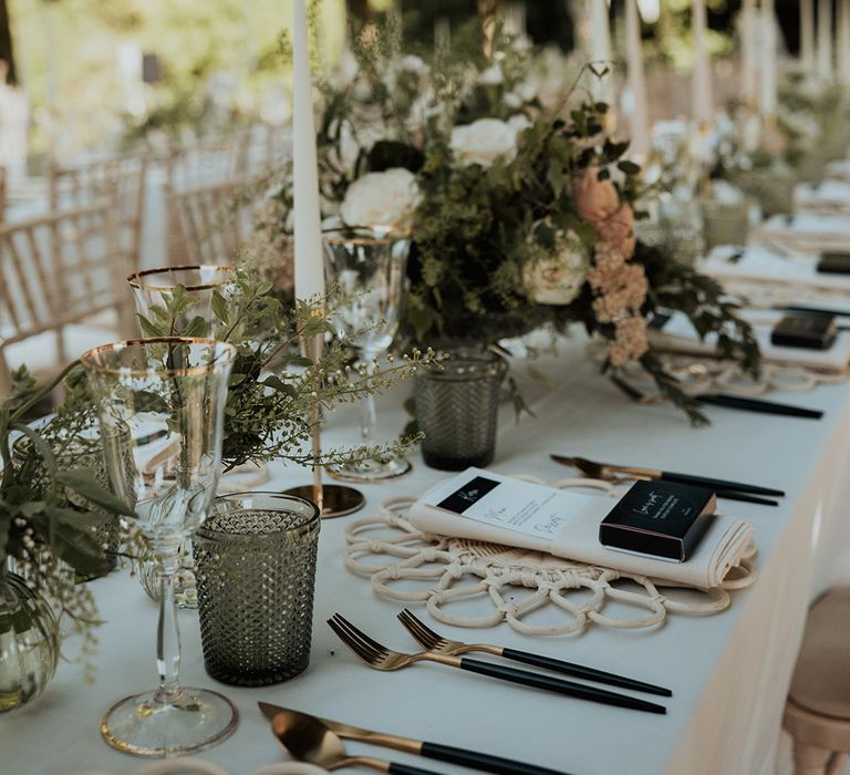 Back and gold cutlery, black and white menu card and white wicker place mats with green glassware, gold rimmed wine glasses and neutral flowers