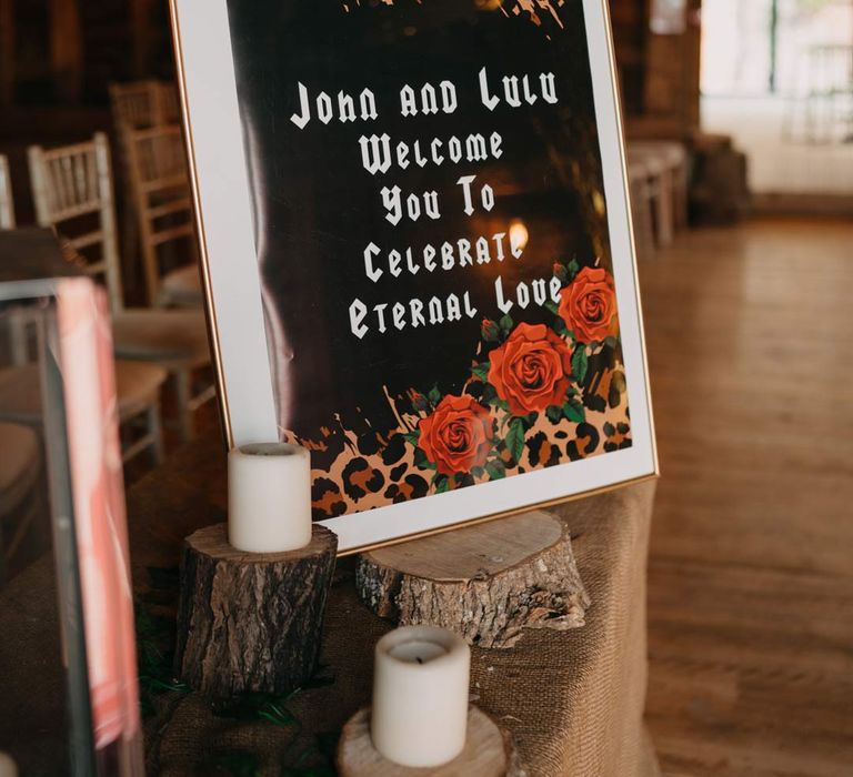 Mirrored wedding welcome sign with red rose and leopard print on wood slice next to pillar candles at Southlands Barn