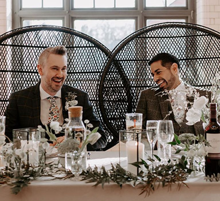 Two groom sit together on peacock chairs at their sweetheart table