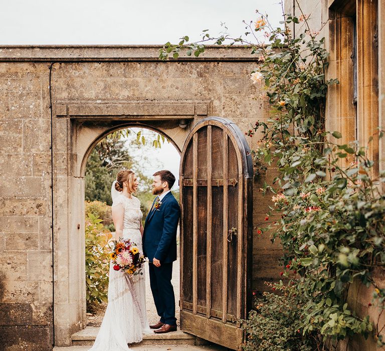 Bride in delicate lace wedding dress with groom in three piece suit posing in doorway at country house 