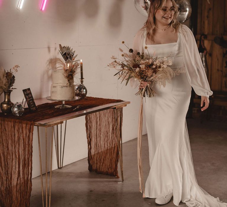 Bride wearing sheer long sleeve wedding dress walking next to the cake table with disco balls and neon signs 