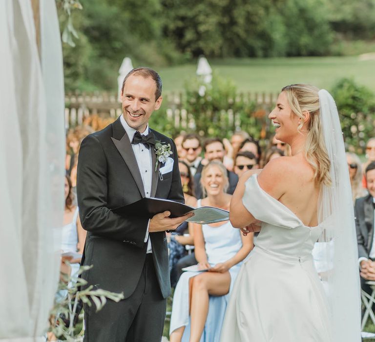 Groom in black tuxedo reading out wedding vows at outdoor wedding ceremony to bride 