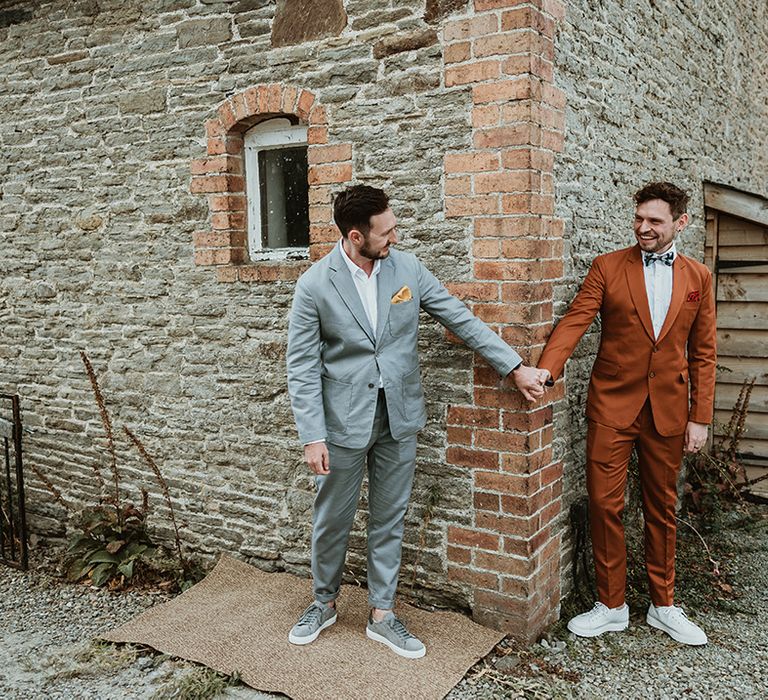 Same sex wedding with two grooms holding hands behind a wall as they see each other for the first time 