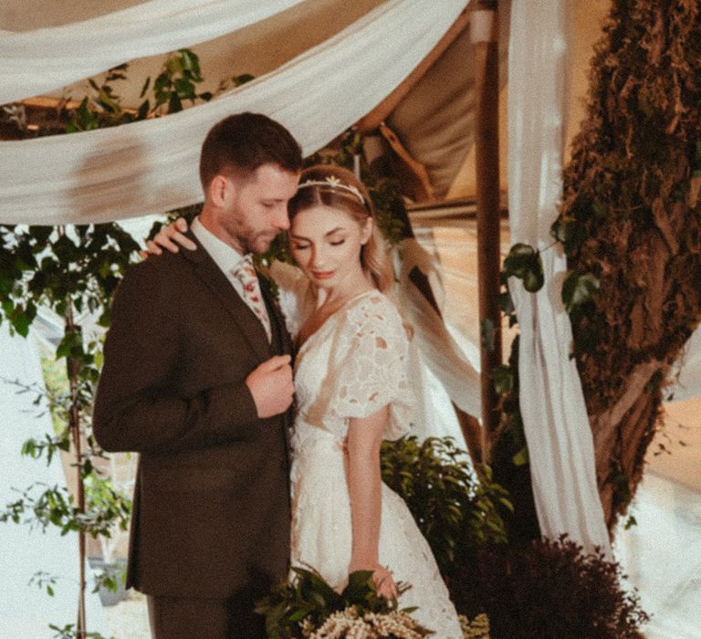 Groom in brown wedding suit with floral tie embracing the bride in a floral lace wedding dress and headband with foliage wedding bouquet