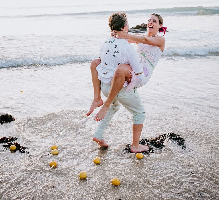 Groom in personalised embroidered waistcoat with the bride in sparkly wedding dress being spun around on the beach 