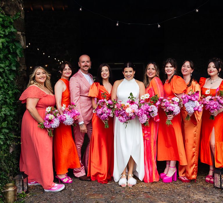 The bridal party wears orange bridesmaid dresses and suits with pink shoes and bouquets for wedding 