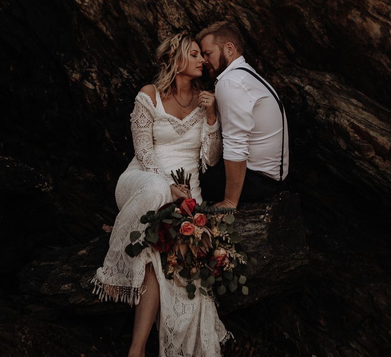 Bride in lace and tassel boho wedding dress with layered jewellery barefoot and large bouquet with groom in shirt and braces 