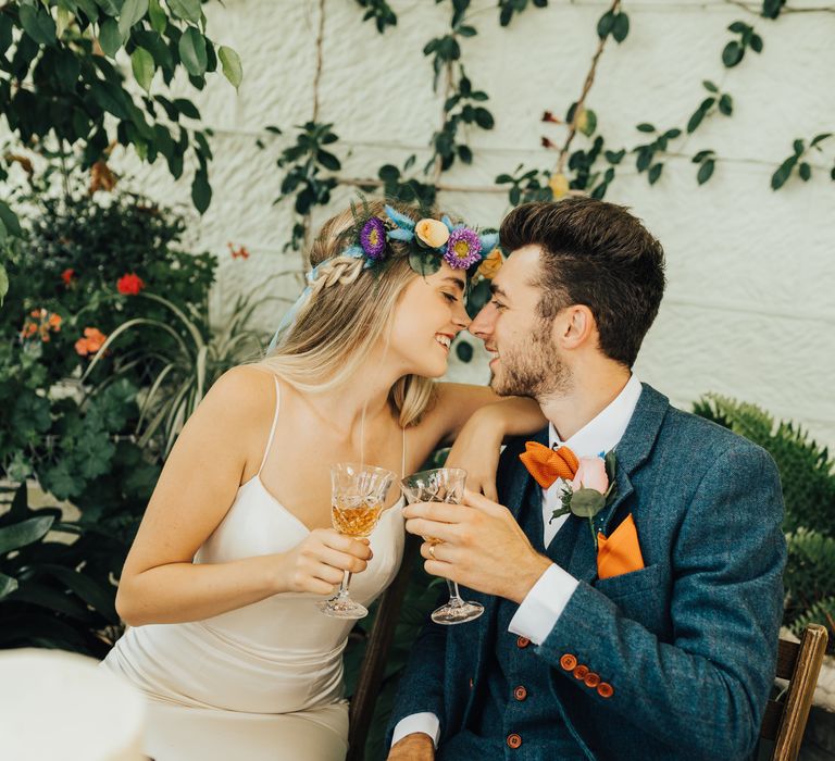 Groom in orange bow tie and bride in colourful flower crown share wine