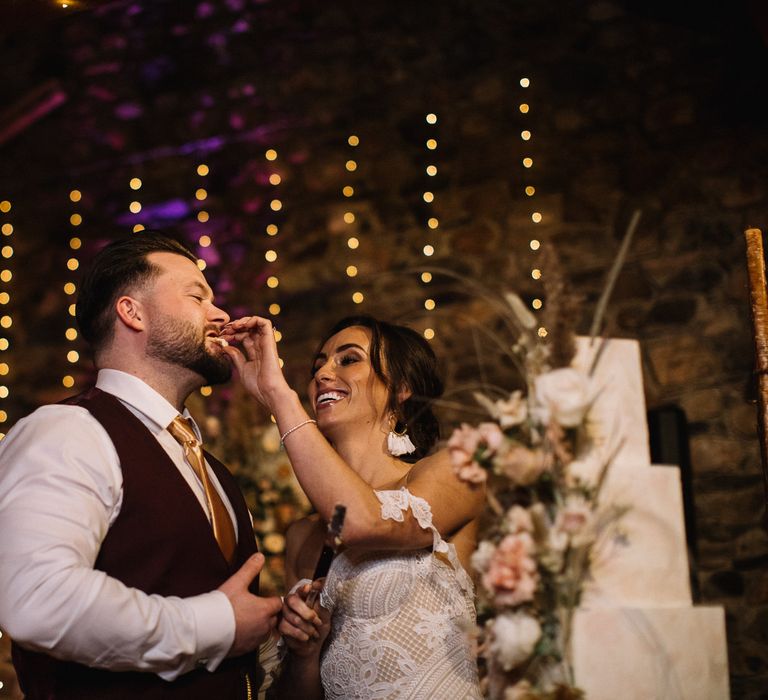 Boho bride feeds groom in burgundy waistcoat wedding cake 