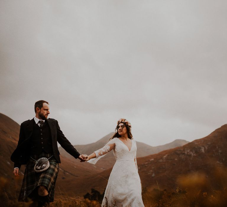 Bride with flower crown and groom in kilt hold hands walking through the Scottish highlands in Glencoe