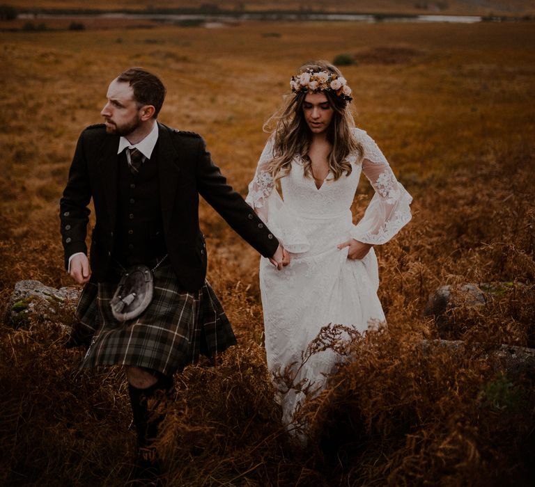 Boho bride in flower crown and groom in kilt walk hand in hand through the highlands in Glencoe