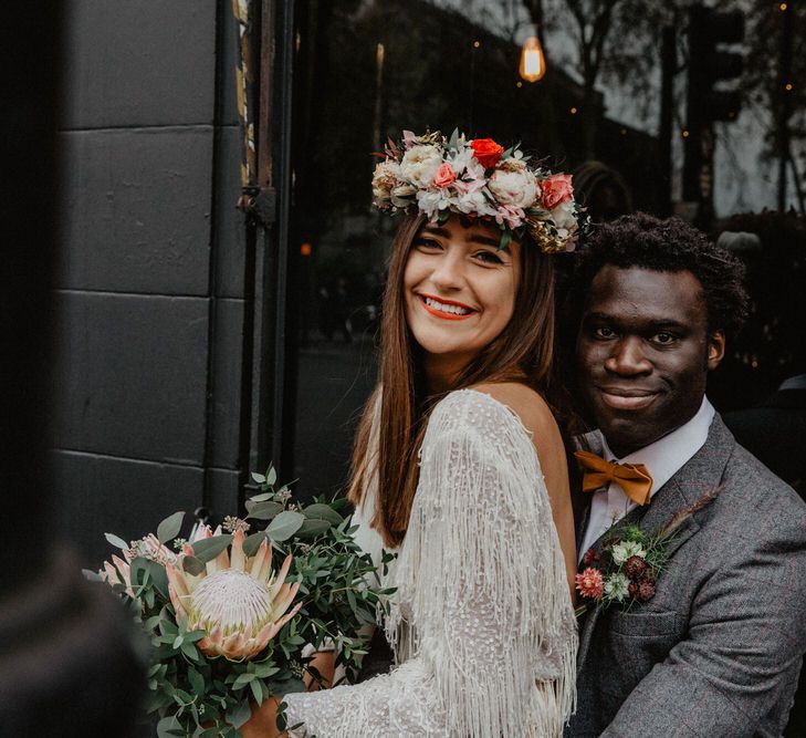 White bride wearing tassel dress and flower crown with protea bouquet sitting on knee of Black groom in tweed suit.