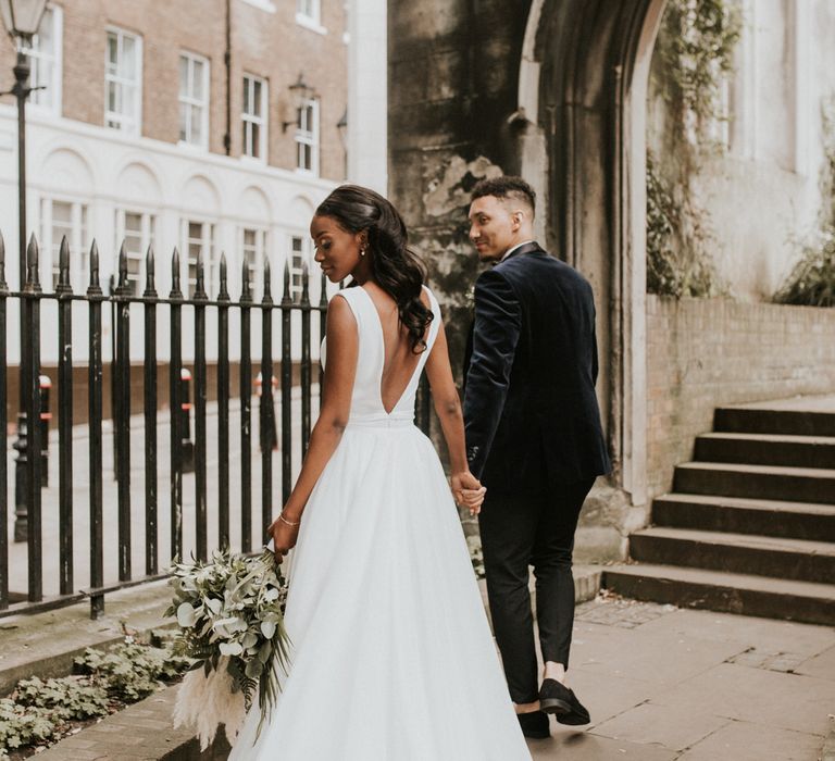 Bride and groom walk away from camera. The bride wears a low back v shaped dress with a long train.