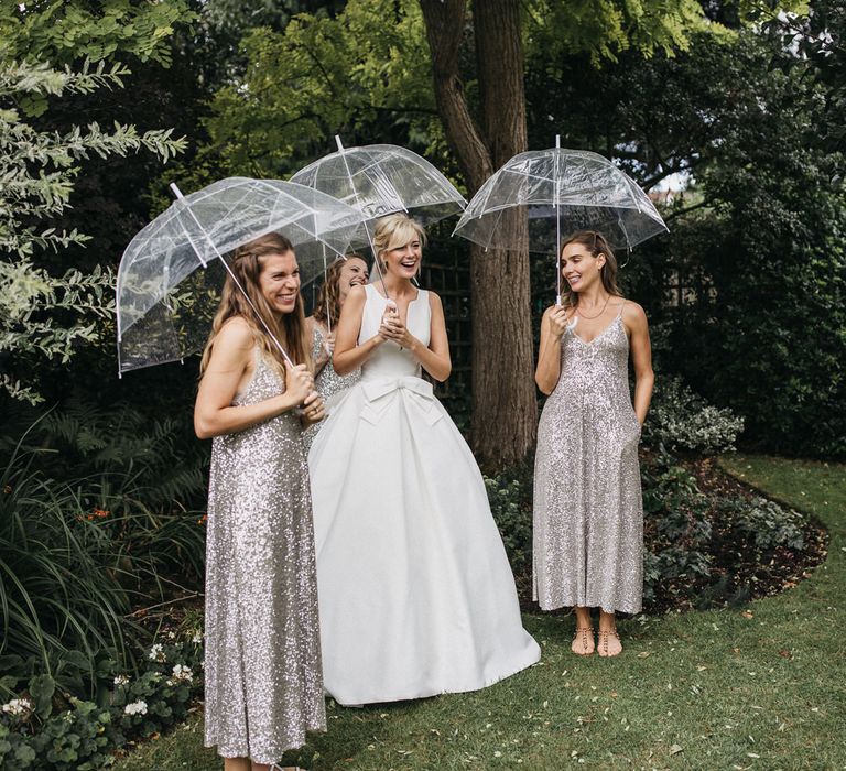 Bride in a Jesus Peiro wedding dress with bow waist detail standing under a clear umbrella with her bridesmaids in silver sequin dresses 