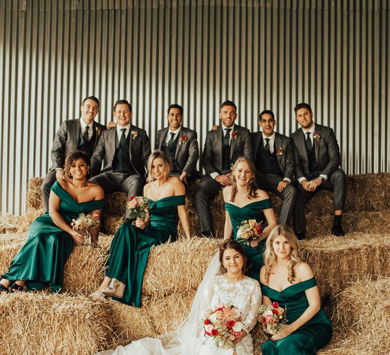 Wedding party portrait with groomsmen in grey wool suits and bridesmaids in dark green off the shoulder dresses sitting on hay bales 