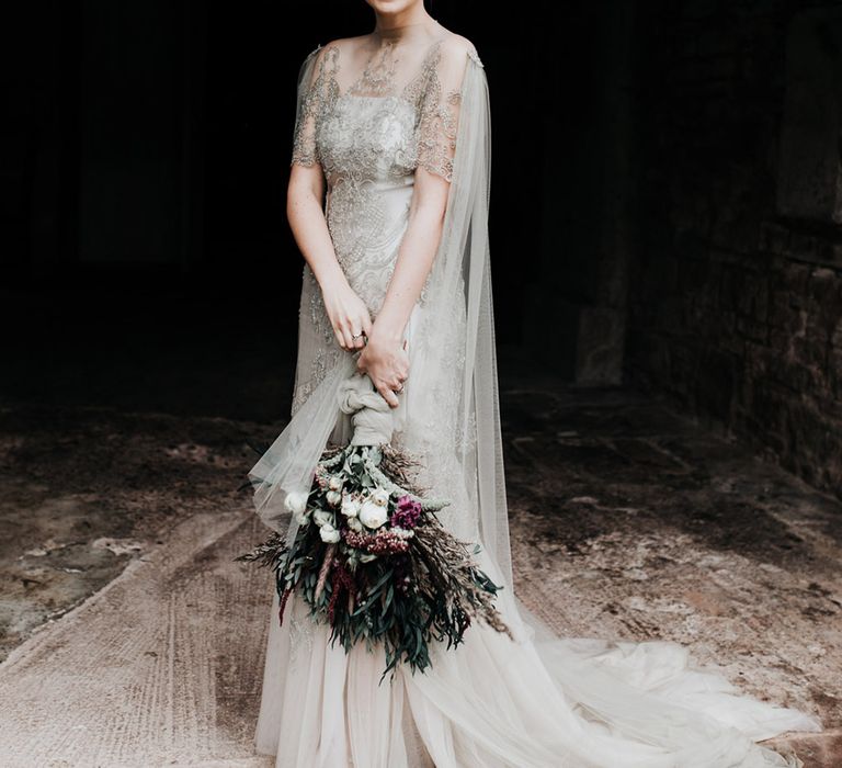 A bride wears a homemade wedding dress with lots of embellishment and a sheer cape. She holds a homemade wedding bouquet.